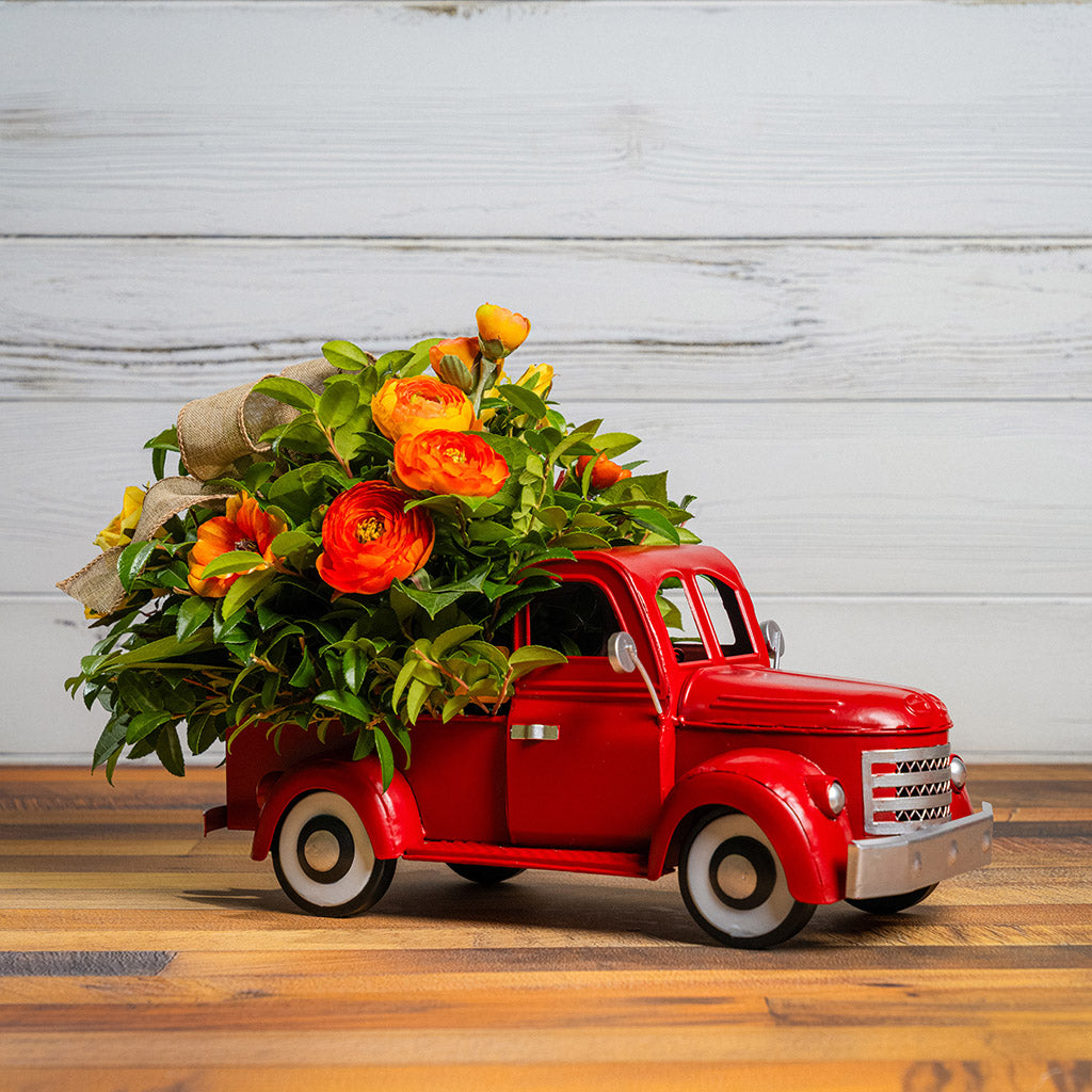 Fresh salal and green huckleberry with faux orange cosmos flowers, faux mini yellow roses, faux orange ranunculus flowers, and a natural linen bow in a red metal truck container