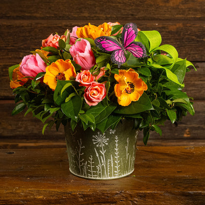 Fresh salal and green huckleberry with faux orange poppies, faux pink tulips, faux peach roses, a bright green bow, and a pink butterfly in a metal green container