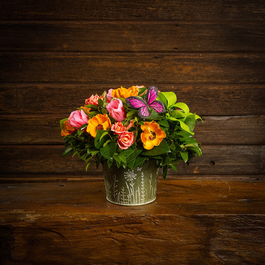 Fresh salal and green huckleberry with faux orange poppies, faux pink tulips, faux peach roses, a bright green bow, and a pink butterfly in a metal green container