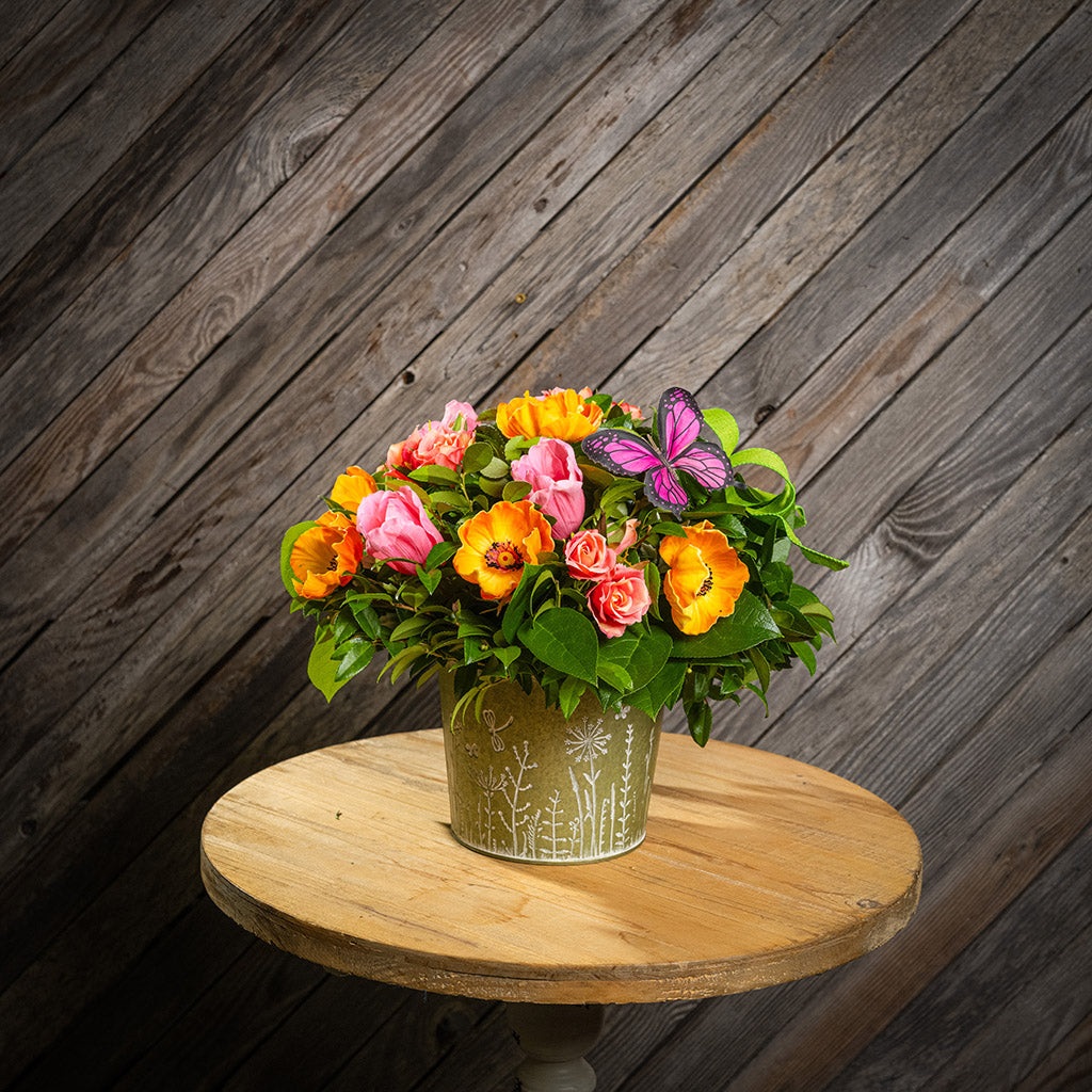 Fresh salal and green huckleberry with faux orange poppies, faux pink tulips, faux peach roses, a bright green bow, and a pink butterfly in a metal green container