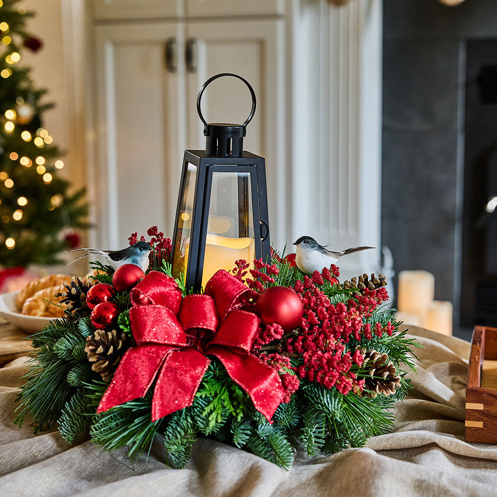 A festive Christmas wreath centerpiece on a beige fabric surface, featuring green pine branches, red berries, a large red bow, pinecones, two small bird figurines, and a black lantern with a lit candle. The background shows a softly blurred indoor holiday scene with a Christmas tree and warm lights.