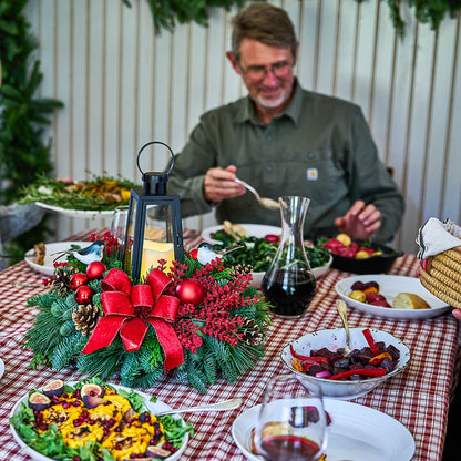 A man in a green shirt sits at a dining table decorated for Christmas, with a red-and-white checkered tablecloth, a holiday wreath centerpiece with a red bow, and plates of food in the foreground.
