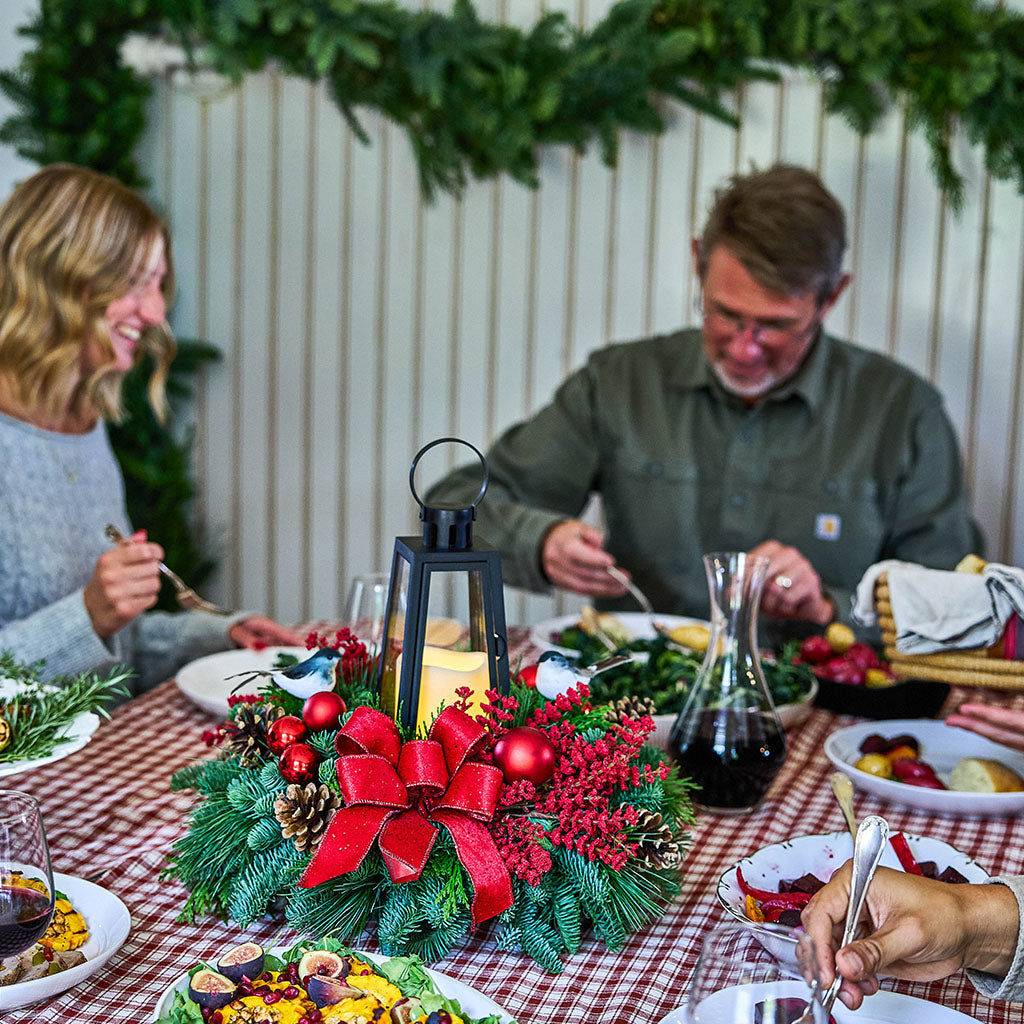 A festive holiday dinner table scene with a man and woman seated at a red-and-white checkered tablecloth, surrounded by food and a central Christmas centerpiece with a large red bow, greenery, pinecones, and red ornaments.
