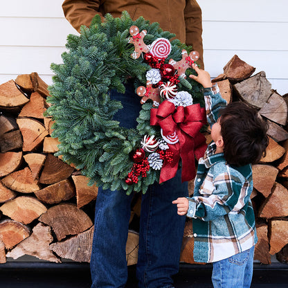 A large green pine wreath decorated with a large red bow, candy canes, gingerbread cookies, and red berries, held by an adult in a brown jacket and jeans while a young child in a green plaid shirt reaches toward it. The background shows stacked firewood against a white siding wall.