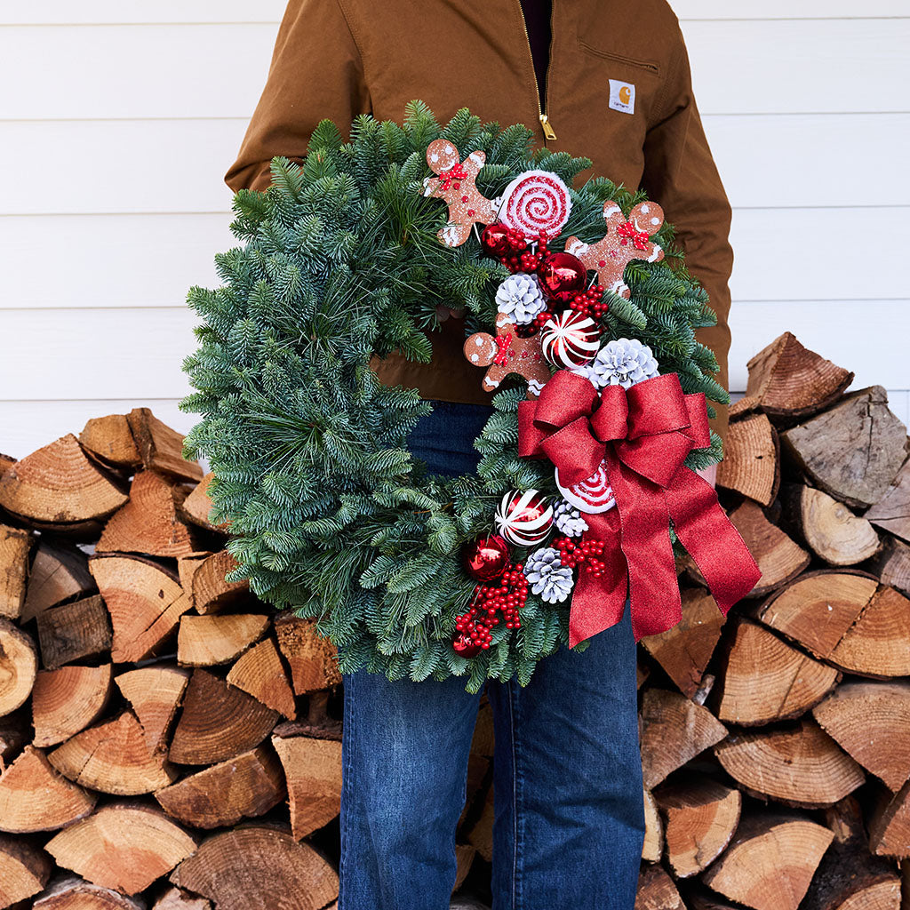 A person in a brown Carhartt jacket and blue jeans holds a green evergreen Christmas wreath decorated with a large red bow, candy canes, gingerbread cookies, and red berries, set against a background of stacked firewood and white siding.