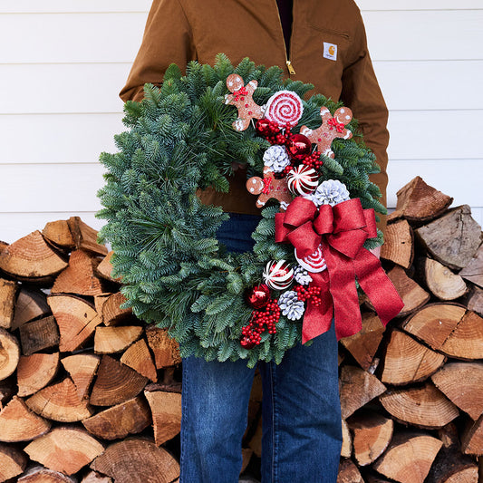 A person in a brown Carhartt jacket and blue jeans holds a green evergreen Christmas wreath decorated with a large red bow, candy canes, gingerbread cookies, and red berries, set against a background of stacked firewood and white siding.