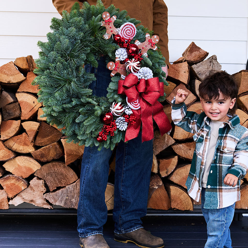 A large green Christmas wreath with red bows, candy canes, pinecones, and red beads is held by an adult standing in front of stacked firewood, with a young child in a plaid jacket holding the adult's hand on the right.