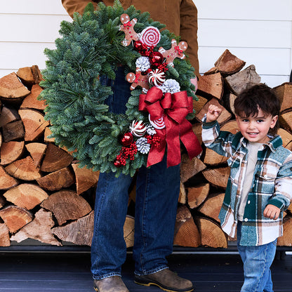 A large green Christmas wreath with red bows, candy canes, pinecones, and red beads is held by an adult standing in front of stacked firewood, with a young child in a plaid jacket holding the adult's hand on the right.