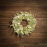 A dried wreath made of natural green hydrangea, cream strawflowers, daisies, cream globe amaranthus, salal leaves or silver-dollar eucalyptus, and sage green eucalyptus hung on a dark wood background.