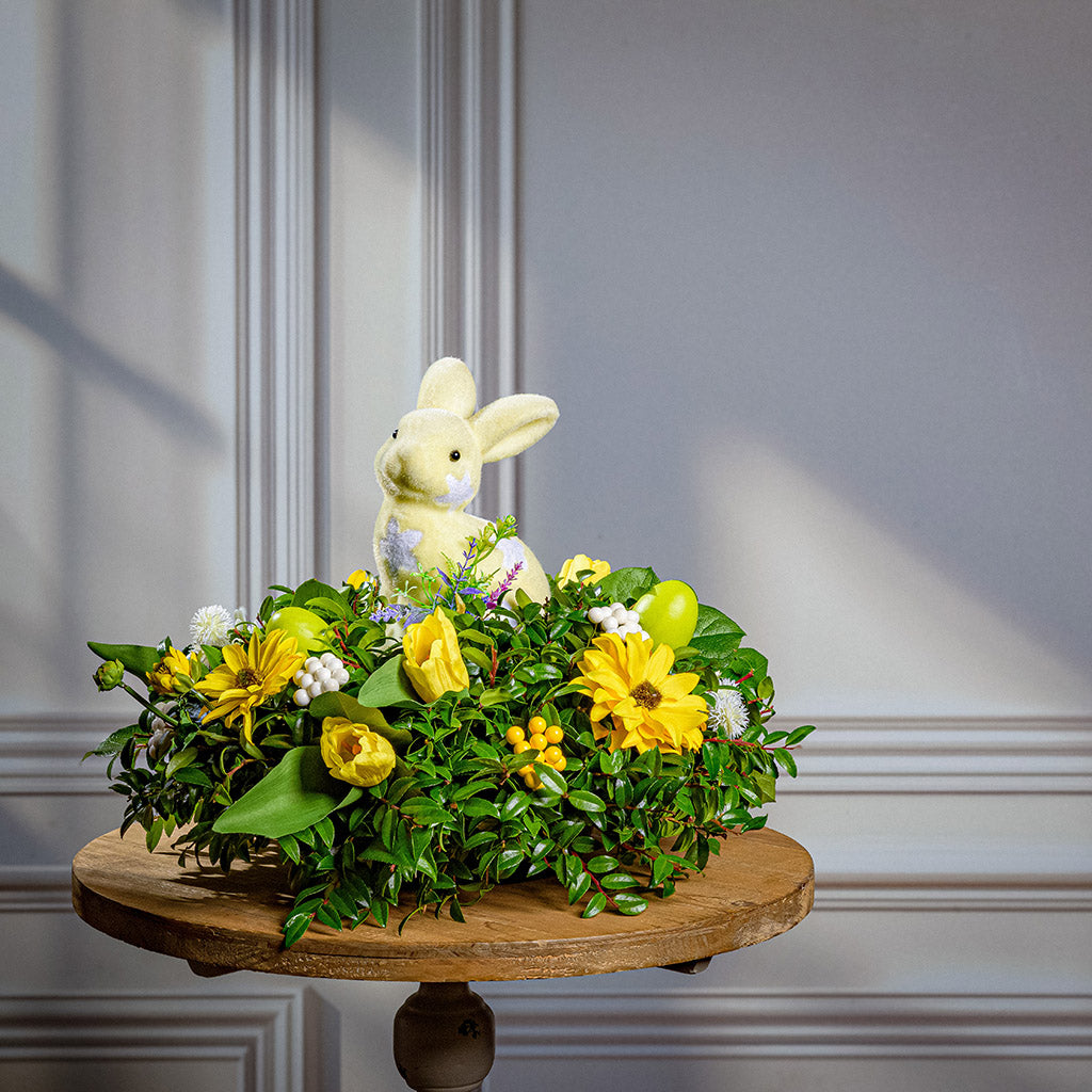 Decorative floral arrangement with a soft bunny on a wooden stand against a gray wall.