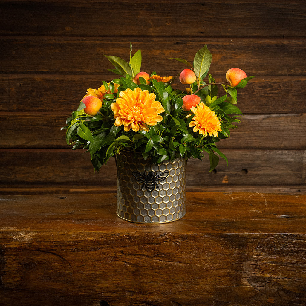 Fresh salal and green huckleberry with faux orange dahlia flowers and peach branches in a metal honeycomb pot with a honey bee