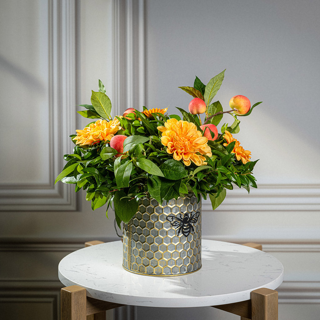 Fresh salal and green huckleberry with faux orange dahlia flowers and peach branches in a metal honeycomb pot with a honey bee