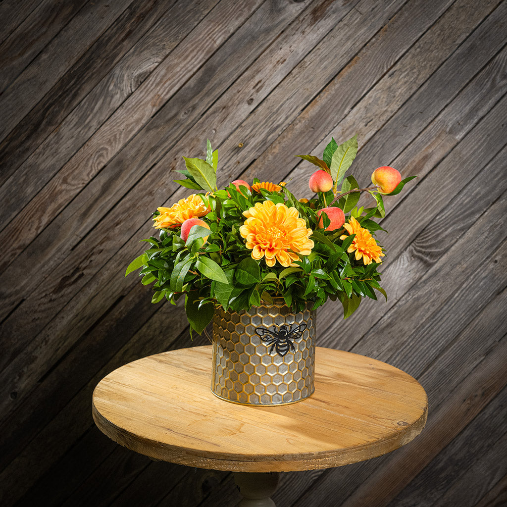 Fresh salal and green huckleberry with faux orange dahlia flowers and peach branches in a metal honeycomb pot with a honey bee