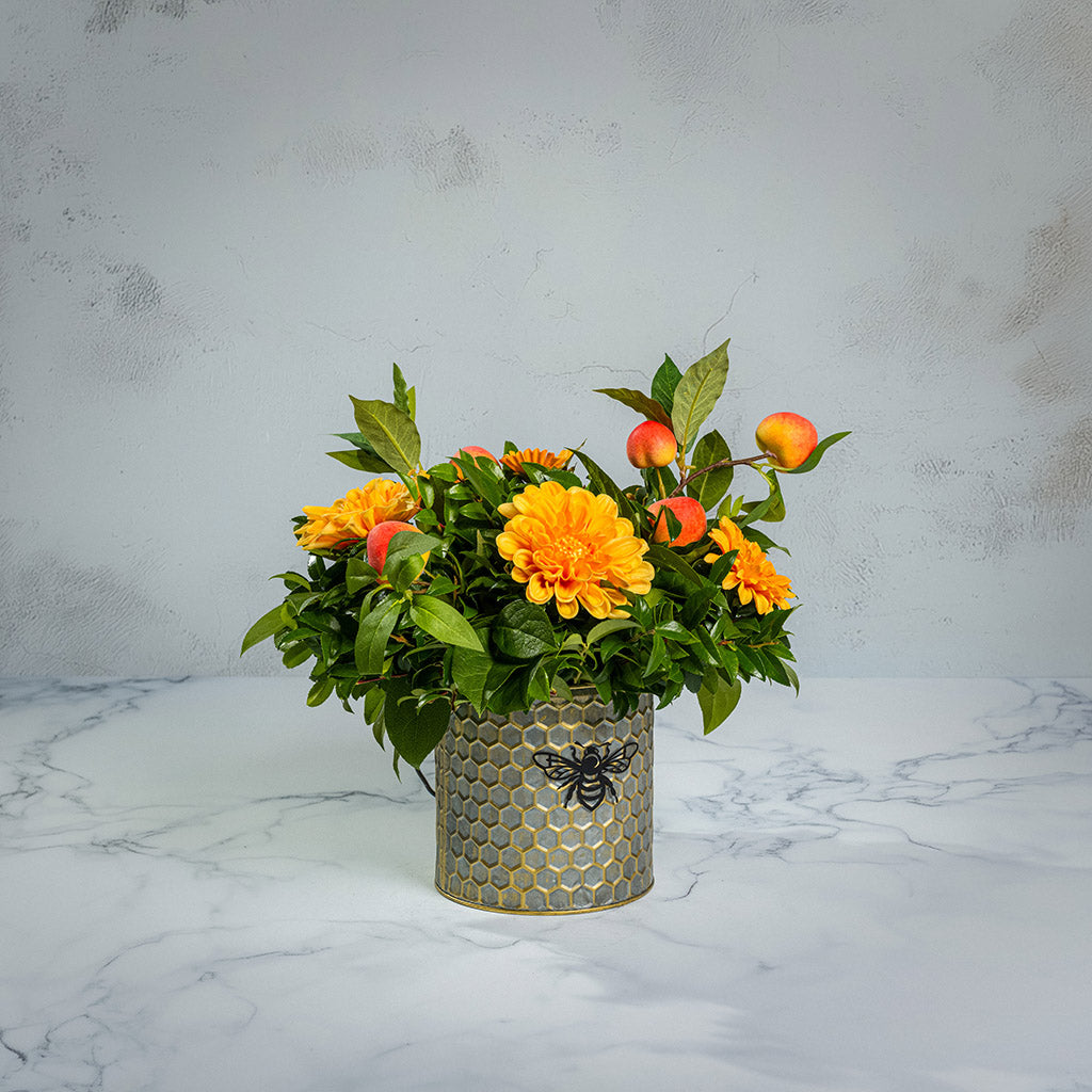 Fresh salal and green huckleberry with faux orange dahlia flowers and peach branches in a metal honeycomb pot with a honey bee