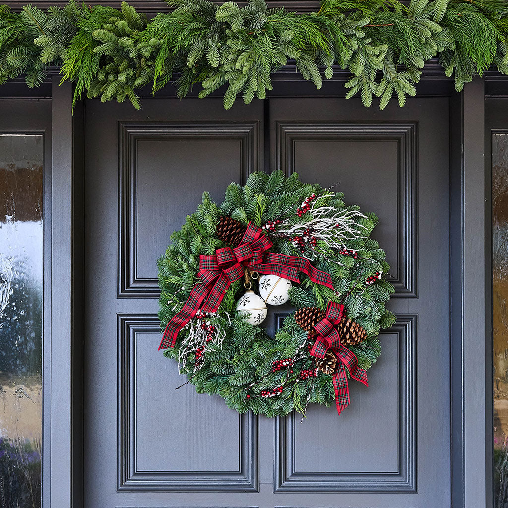 A festive Christmas wreath with green pine branches, red plaid ribbons, white ornaments, and pinecones hanging on a dark gray front door, with a matching green garland across the top.
