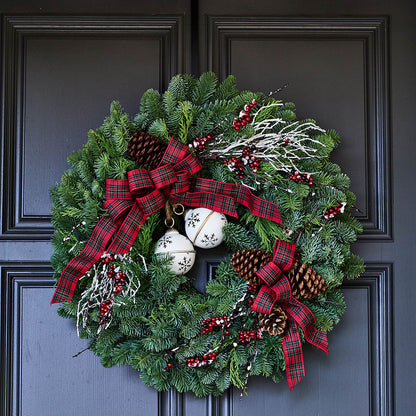 A festive Christmas wreath made of green pine branches, decorated with red and black plaid ribbons, white bells with black snowflakes, pinecones, and red berries, hanging on a dark charcoal paneled door.