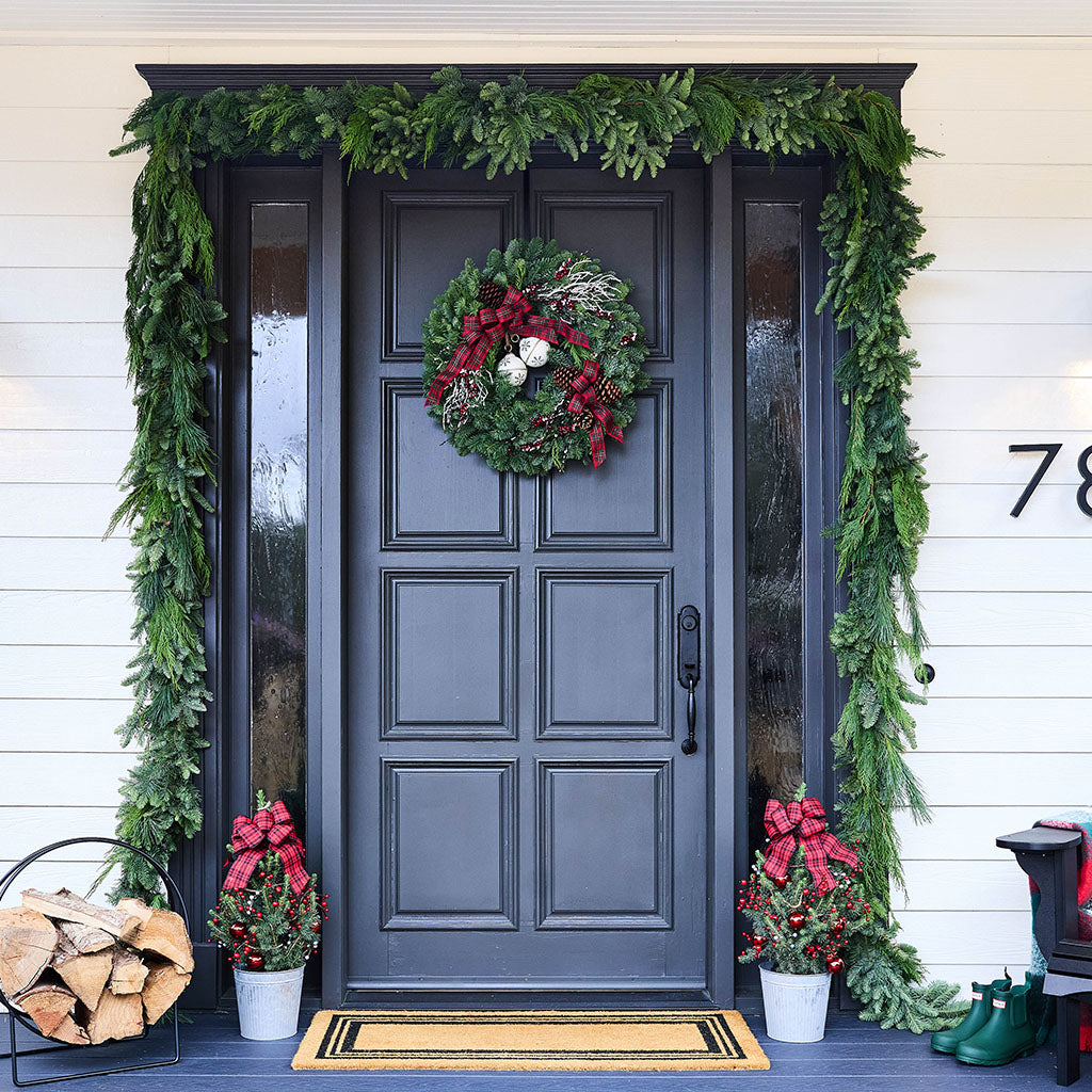 A front door decorated for Christmas, featuring a dark charcoal gray door with a green pine garland framing the entryway, a matching wreath with red berries and pinecones on the door, potted holiday plants with red bows on the porch, and a log basket beside the door.