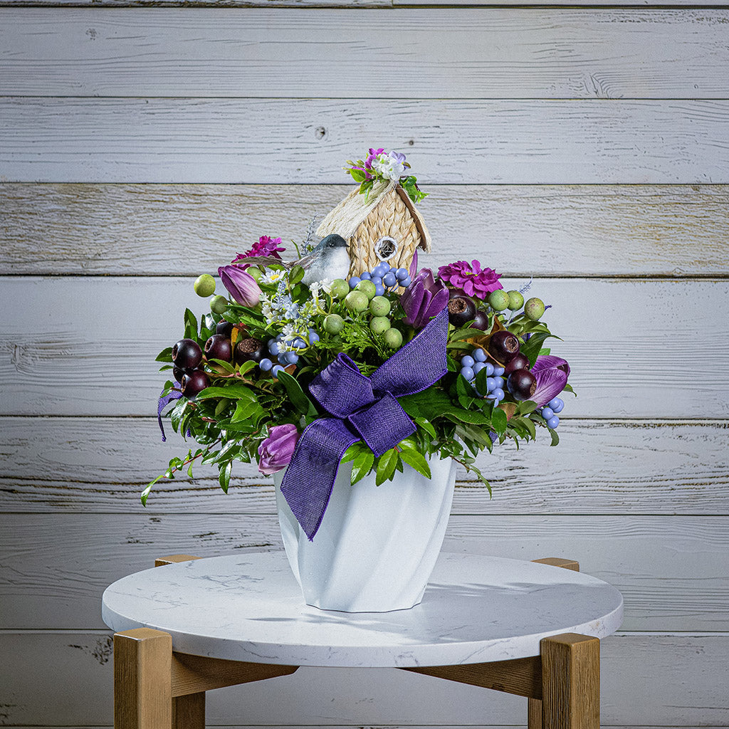 Floral arrangement with a birdhouse and purple bow on a marble table against a wooden background