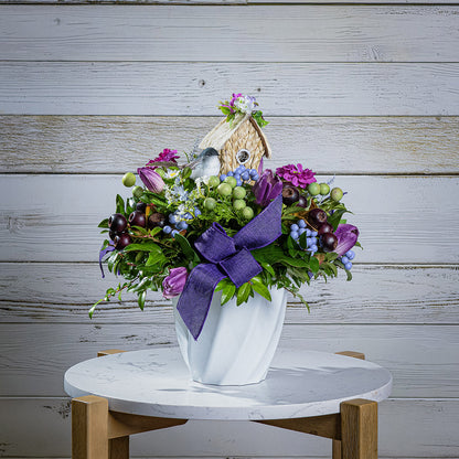Floral arrangement with a birdhouse and purple bow on a marble table against a wooden background
