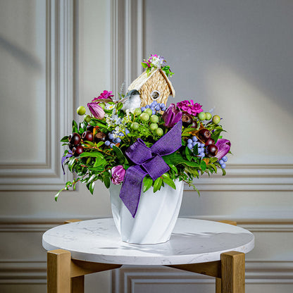Floral arrangement with a birdhouse and purple ribbon on a marble table.