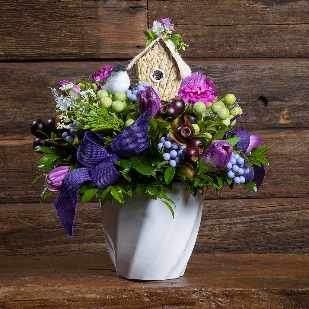 Floral arrangement with a birdhouse and purple accents in a white vase on a wooden surface.
