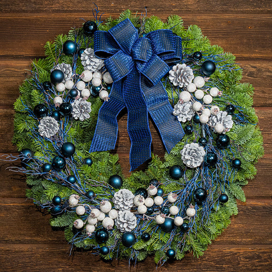 A festive Christmas wreath made of green pine branches, decorated with a large blue patterned bow, white berries, white pinecones, and dark blue ornaments, displayed on a dark brown wooden background.