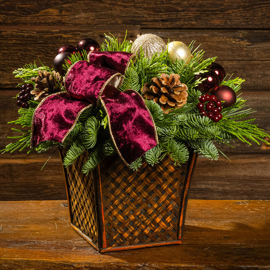noble fir, cedar, and pine with burgundy and gold ball ornaments, faux burgundy berries, natural pinecones, and a burgundy velvet bow in a brown wooden container