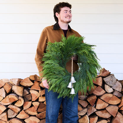 A man wearing a brown jacket and blue jeans stands in front of a white siding wall and a stacked firewood pile, holding a green evergreen wreath with small silver bells.