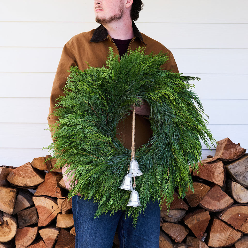 A man wearing a brown jacket and blue jeans holds a green evergreen wreath with three hanging silver bells, standing in front of a white wood-paneled wall and a stacked firewood pile.