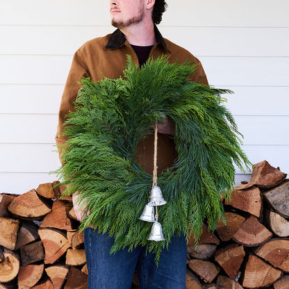 A man wearing a brown jacket and blue jeans holds a green evergreen wreath with three hanging silver bells, standing in front of a white wood-paneled wall and a stacked firewood pile.