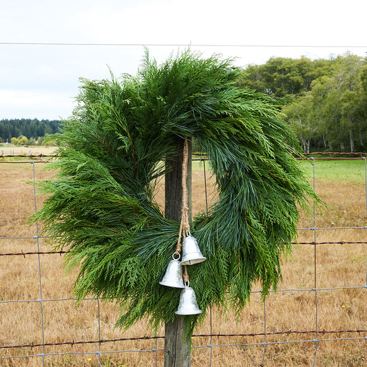 A green evergreen wreath hanging on a rustic wooden post in front of a wire fence, with three small silver bells dangling from the center and an outdoor field and trees in the background.