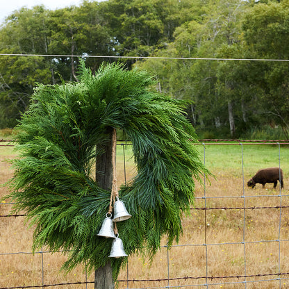 A green evergreen wreath with three hanging silver bells is attached to a wooden post in a wire fence, with a grassy field and trees in the background.