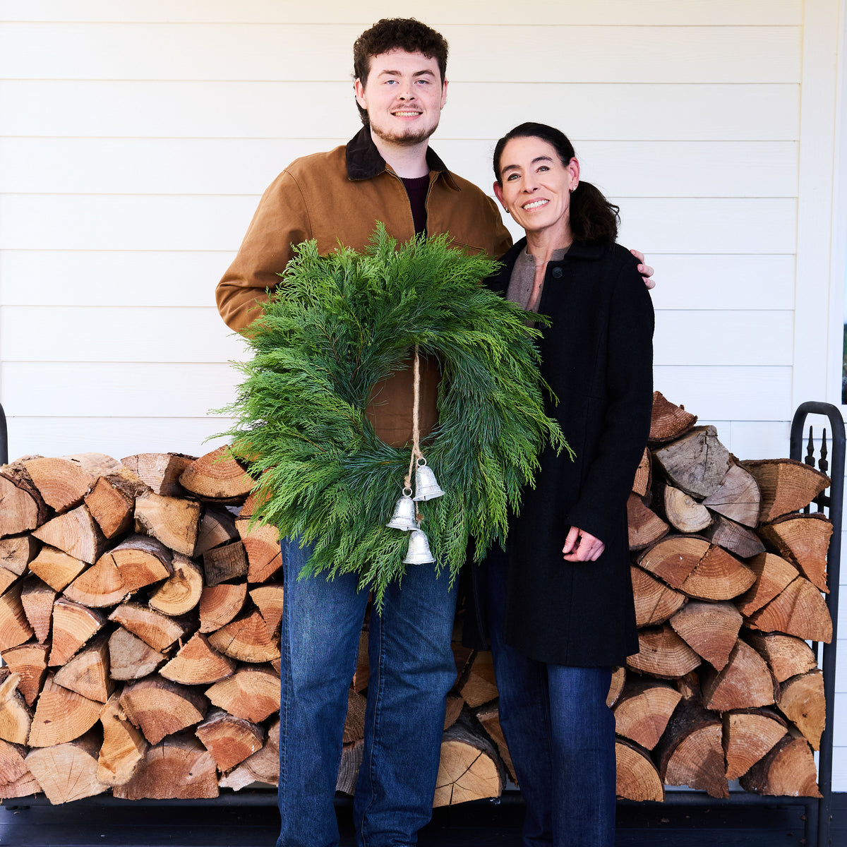 A man and a woman standing together in front of a white siding wall and a stacked firewood pile, holding a green evergreen wreath with small white bells.