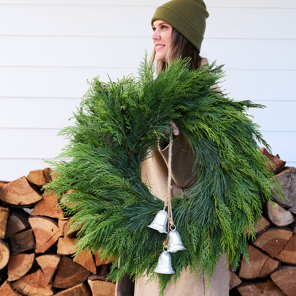A woman wearing an olive green beanie and a brown coat holds a large green evergreen wreath decorated with three silver hanging bells. She is standing in front of a white paneled wall and a stacked woodpile.