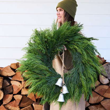 A woman wearing an olive green beanie and a brown coat holds a large green evergreen wreath decorated with three silver hanging bells. She is standing in front of a white paneled wall and a stacked woodpile.