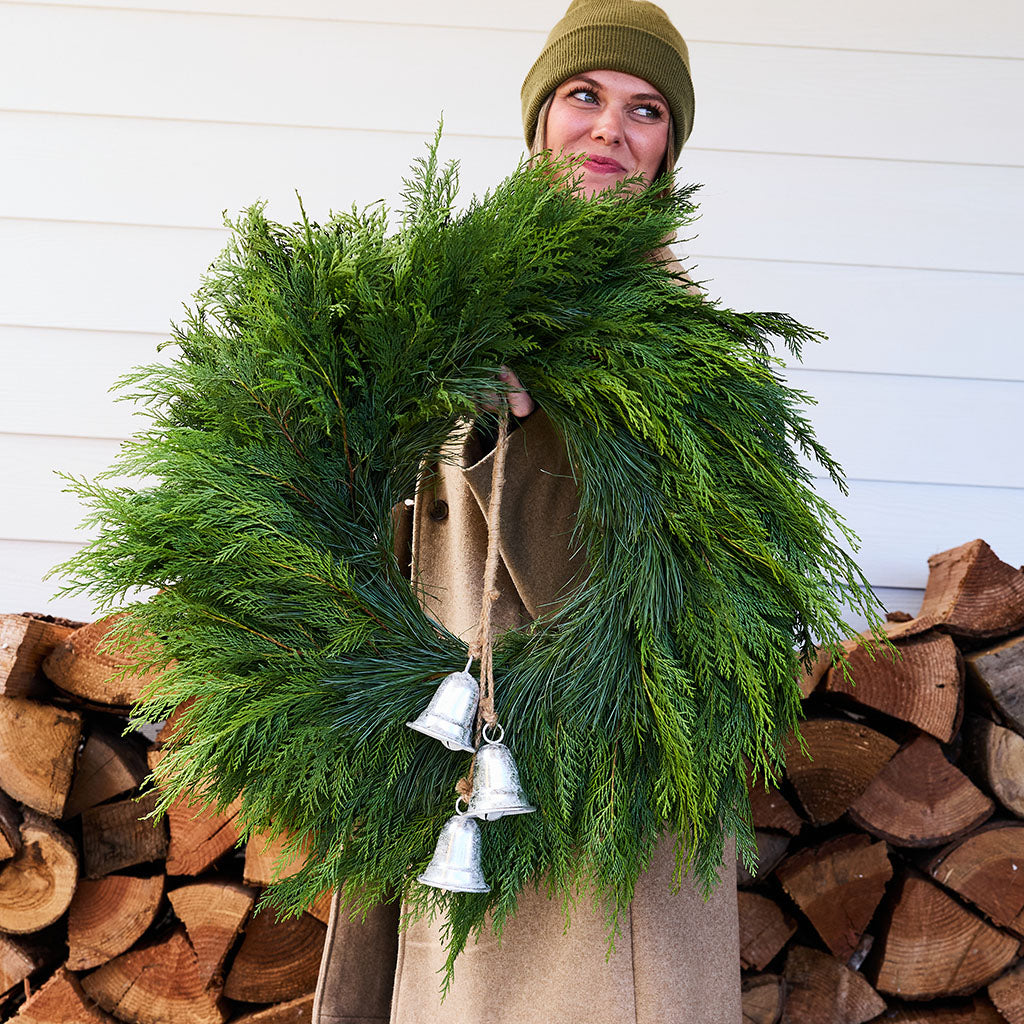 A person wearing a green beanie and tan coat holds a large green evergreen wreath decorated with three silver hanging bells, standing in front of a white siding wall and stacked firewood.