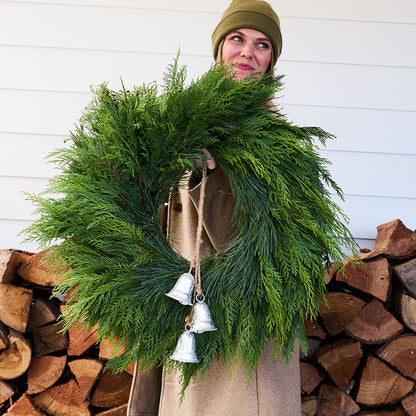 A person wearing a green beanie and tan coat holds a large green evergreen wreath decorated with three silver hanging bells, standing in front of a white siding wall and stacked firewood.