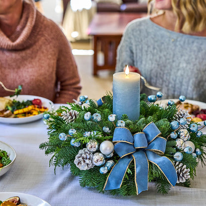 A lit blue pillar candle sits in a festive evergreen wreath centerpiece on a dining table, decorated with blue and silver ornaments, pinecones, and a large blue ribbon bow. Two people are seated at the head of the table in a warmly lit indoor setting.