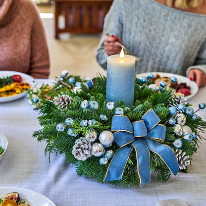 A festive Christmas wreath centerpiece on a white tablecloth, featuring green pine branches, blue ribbon bow, white and blue ornaments, pinecones, and a lit blue pillar candle. Parts of two people at the head of the table are visible in the background.