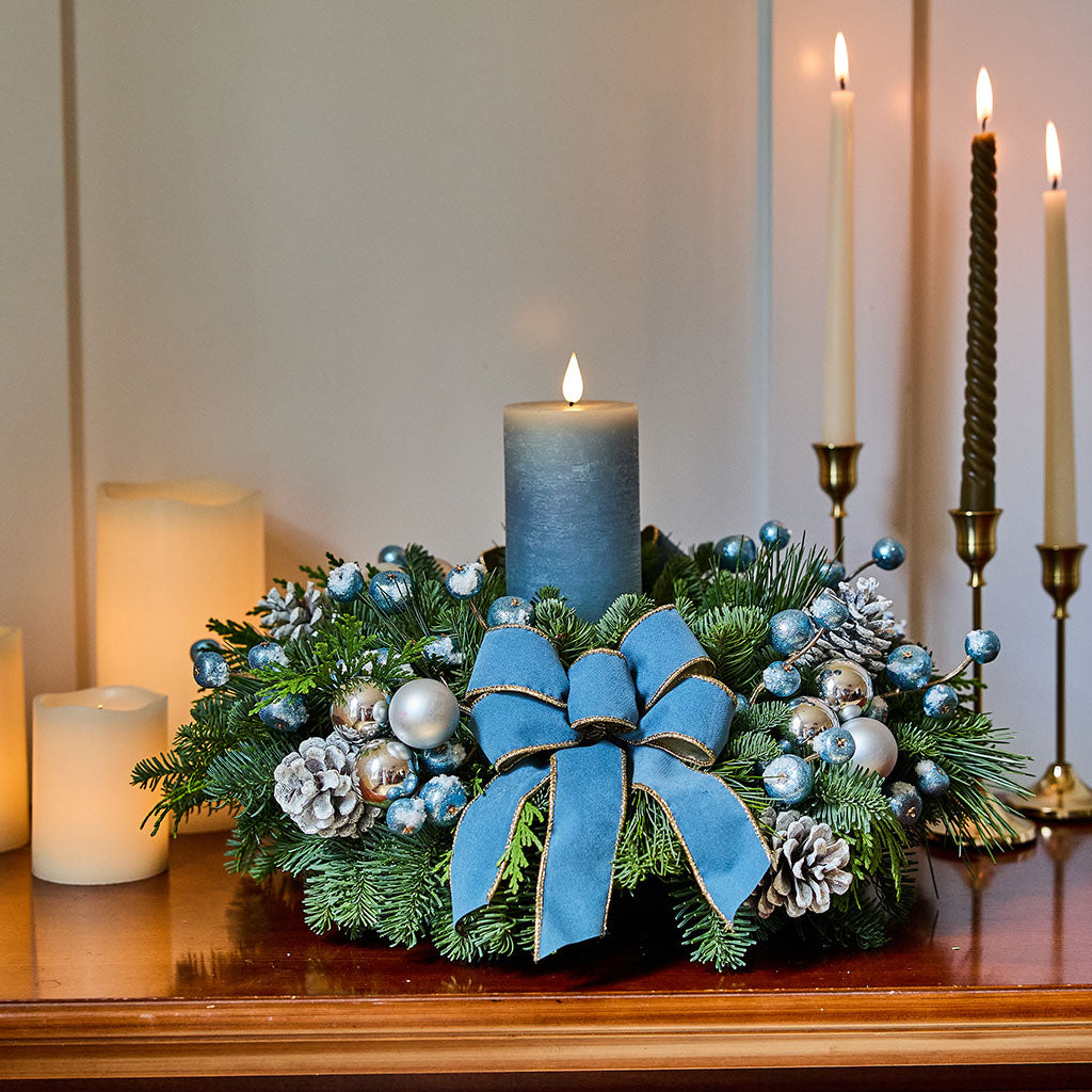 A festive Christmas wreath centerpiece on a wooden mantel, featuring green pine branches, blue and silver ornaments, pinecones, and a large blue velvet bow, with lit candles in the background.