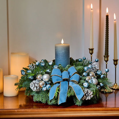 A festive Christmas wreath centerpiece on a wooden mantel, featuring green pine branches, blue and silver ornaments, pinecones, and a large blue velvet bow, with lit candles in the background.