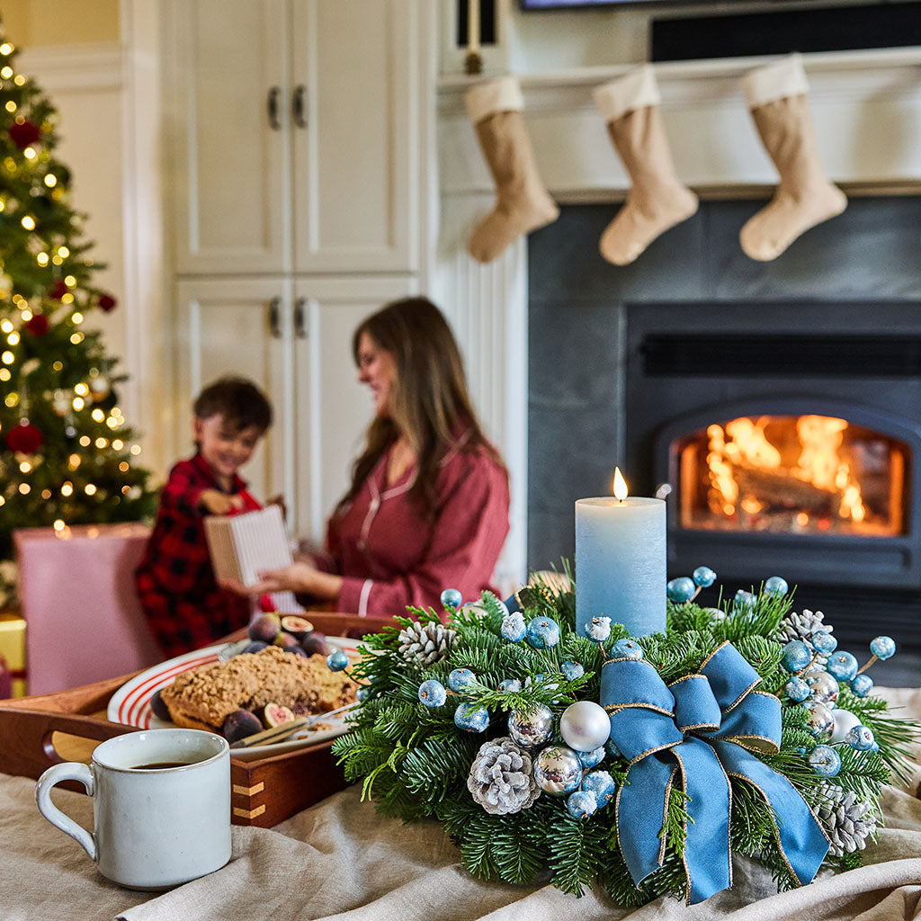 A festive Christmas wreath centerpiece on a wooden mantel, featuring green pine branches, blue and silver ornaments, pinecones, and a large blue velvet bow, with lit candles in the background.