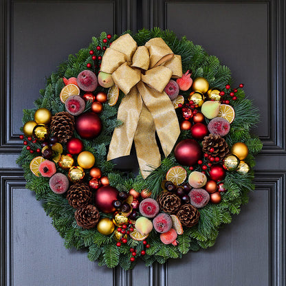 A festive Christmas wreath with a large gold bow, decorated with red and gold ornaments, pinecones, dried fruit, and red berries, hung on a dark gray paneled door.