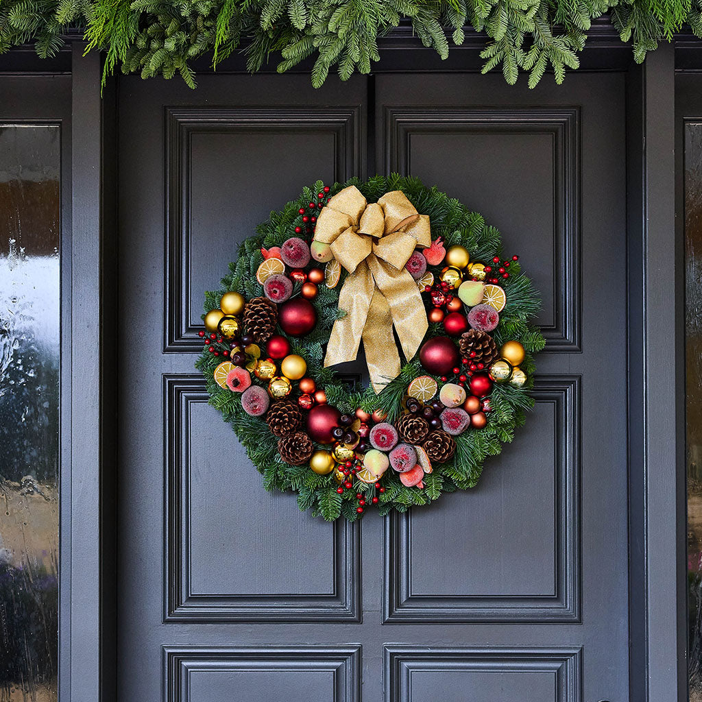A festive Christmas wreath hanging on a dark charcoal front door, decorated with green pine branches, a large gold ribbon bow, red and gold ornaments, pinecones, berries, and small pink and yellow accents.