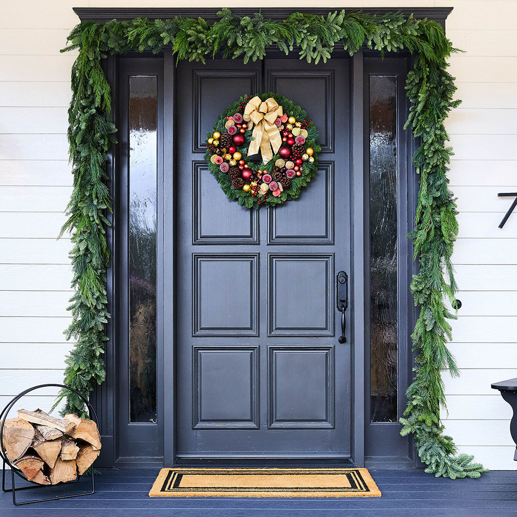 A festive holiday front door decorated with a green pine garland, a matching wreath with red ornaments and a large gold bow, a black paneled door, white siding, a wood log basket, and a doormat.