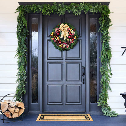 A festive holiday front door decorated with a green pine garland, a matching wreath with red ornaments and a large gold bow, a black paneled door, white siding, a wood log basket, and a doormat.