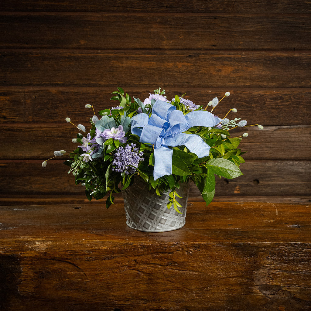 Fresh salal and green huckleberry with faux lilac delphinium flowers, faux purple verbena, faux pussy willows, and a periwinkle bow in a metal white patterned pot