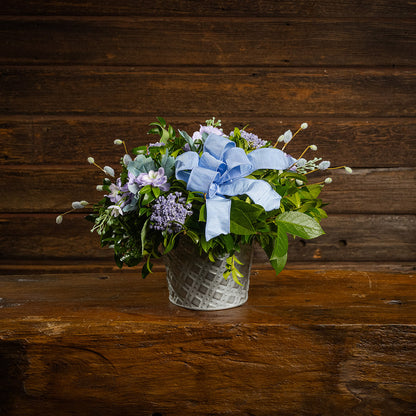 Fresh salal and green huckleberry with faux lilac delphinium flowers, faux purple verbena, faux pussy willows, and a periwinkle bow in a metal white patterned pot