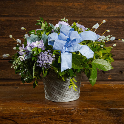 Fresh salal and green huckleberry with faux lilac delphinium flowers, faux purple verbena, faux pussy willows, and a periwinkle bow in a metal white patterned pot