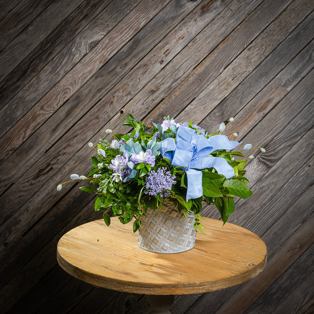 Fresh salal and green huckleberry with faux lilac delphinium flowers, faux purple verbena, faux pussy willows, and a periwinkle bow in a metal white patterned pot